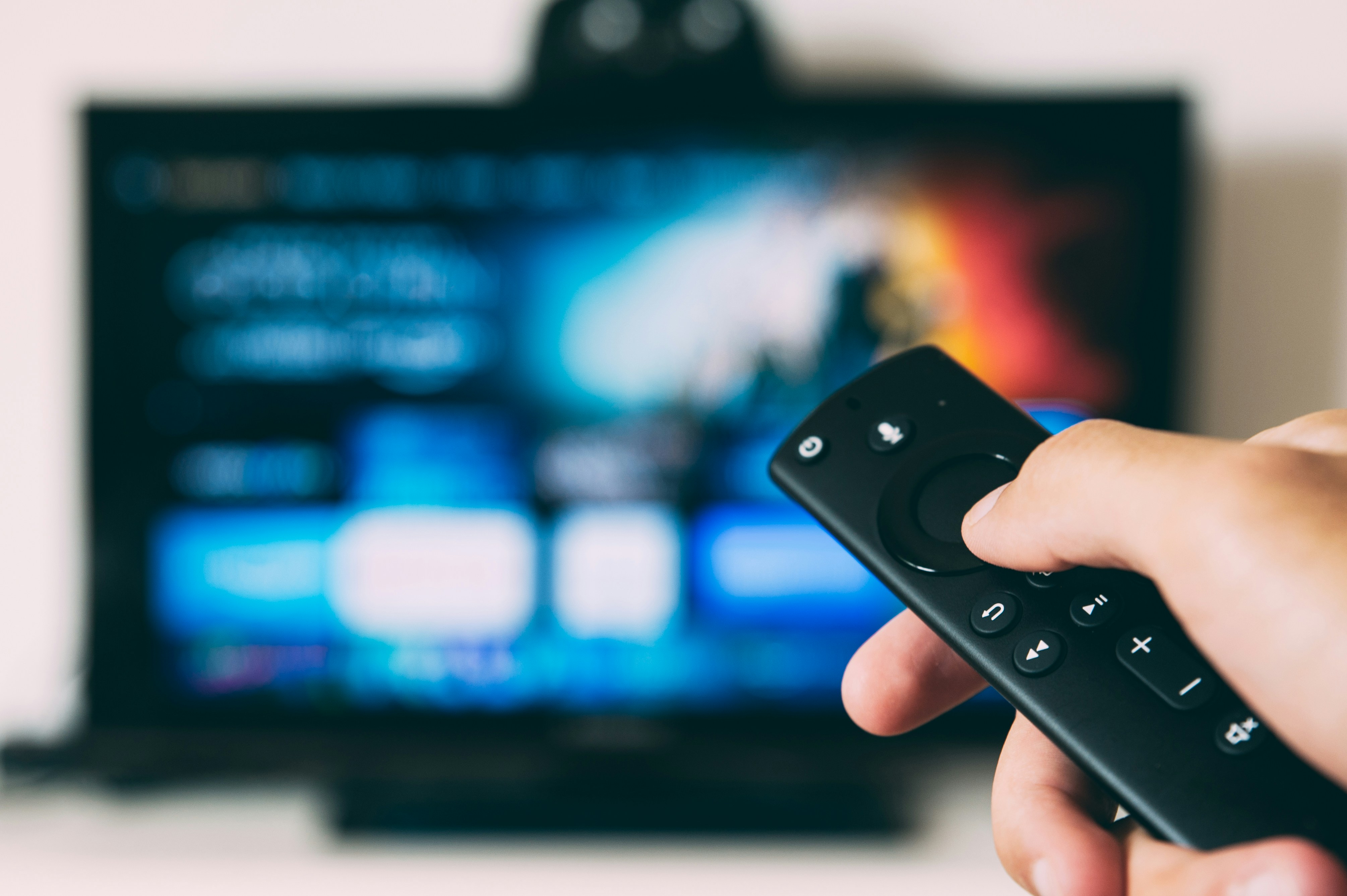 A hand holding a television remote control pointed toward a turned-on flat screen TV.