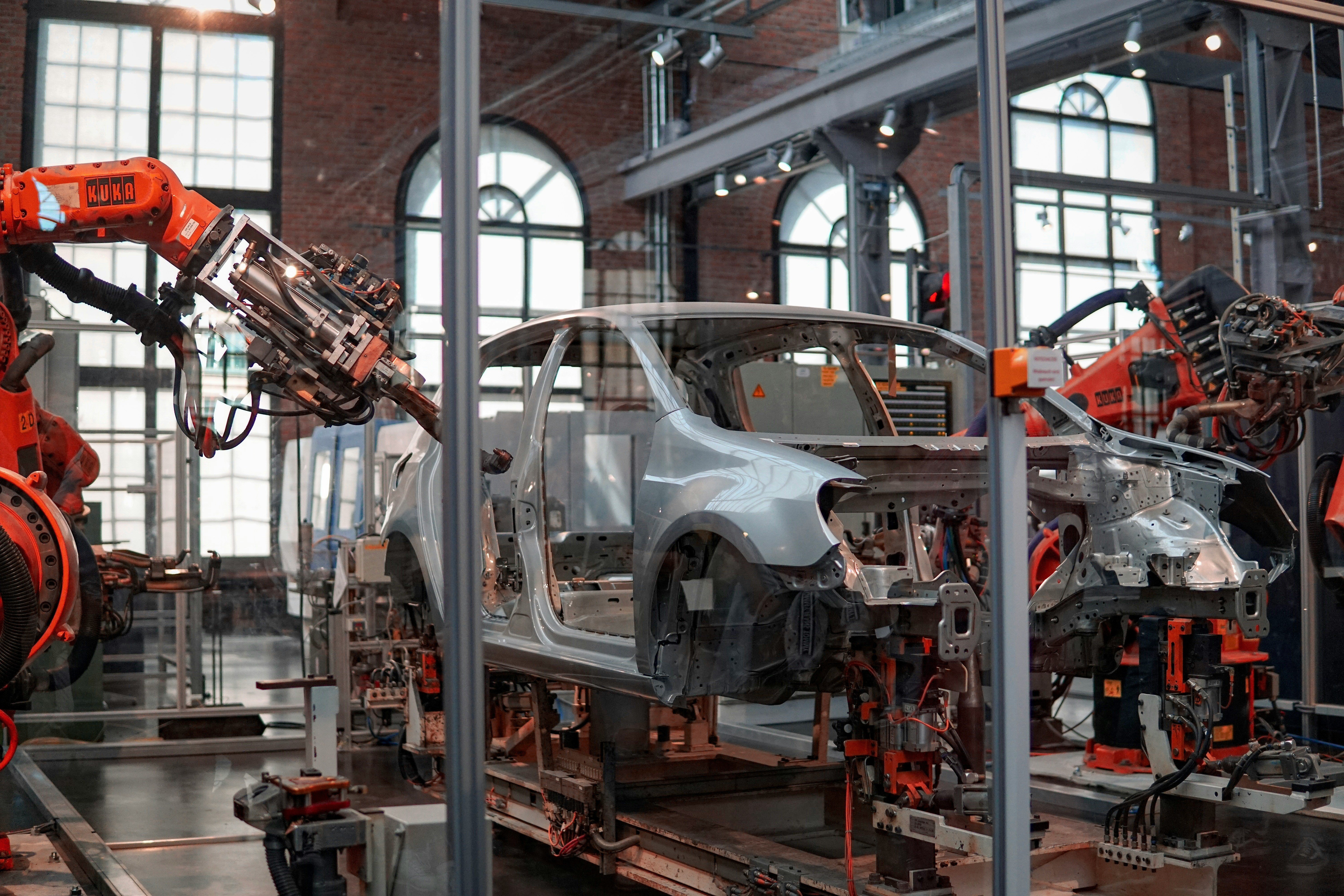 Two industrial robots work on a partially assembled silver car body inside an automotive factory.