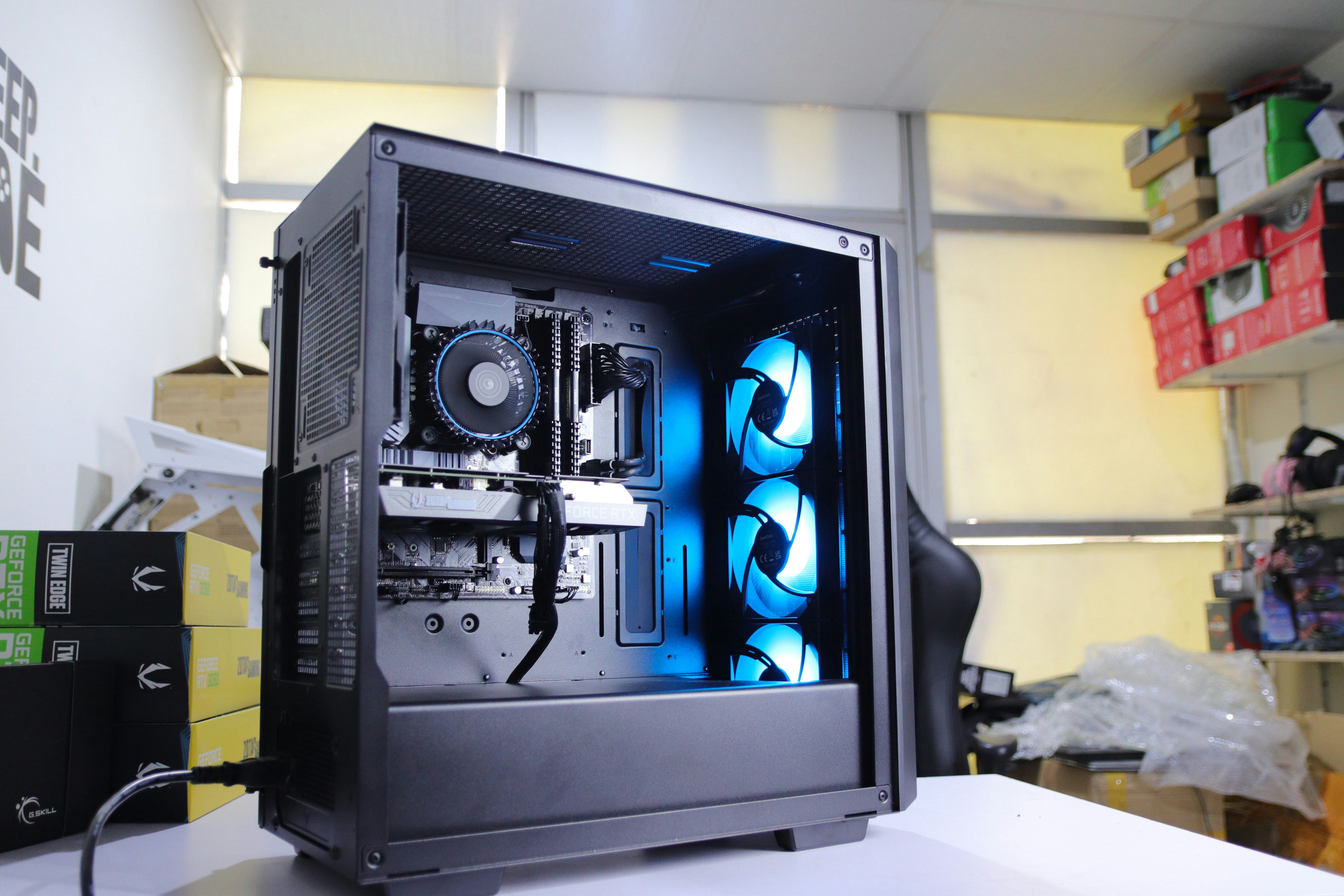 An open black desktop computer case with an RTX graphics card and three blue-lit intake fans, sitting on a workbench.