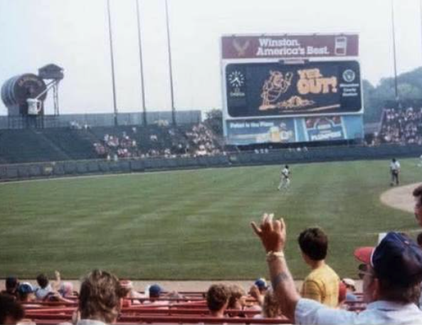 A view across a mostly empty ballpark toward an outfield scoreboard with a cartoon mascot and the words YER OUT, with a fan's raised arm and a few spectators in the foreground and thin crowds scattered through the outfield stands.
