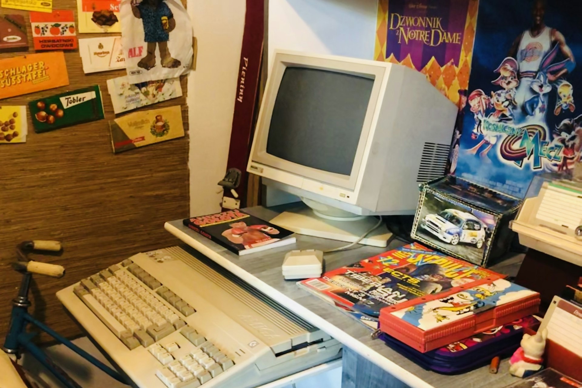 A teenager's bedroom computer desk from the 1990s, with a beige keyboard computer and CRT monitor, a stack of gaming magazines, and a Looney Tunes pencil case, next to a wall of chocolate bar wrappers and Polish posters for The Hunchback of Notre Dame and Space Jam.